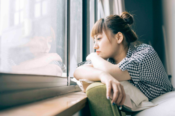 A woman sits, looking out of a windowsill with her chin resting on her folded arms